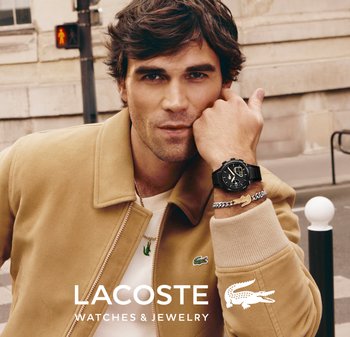 Hombre con chaqueta beige mostrando un reloj de pulsera negro y una pulsera plateada con el logotipo de un cocodrilo, de pie en una esquina de calle urbana.
