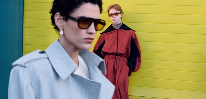 To modebevidste personer i et rum: den ene justerer en højhælet sko, mens hun sidder, den anden poserer iført solbriller, bluse og mønstret nederdel.