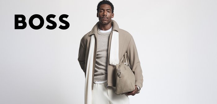 Giacca beige chiaro con colletto alto, indossata sopra un maglione grigio, abbinata a pantaloni bianchi. Tiene una borsa morbida, color marrone chiaro. Testo nero "BOSS" sulla sinistra.