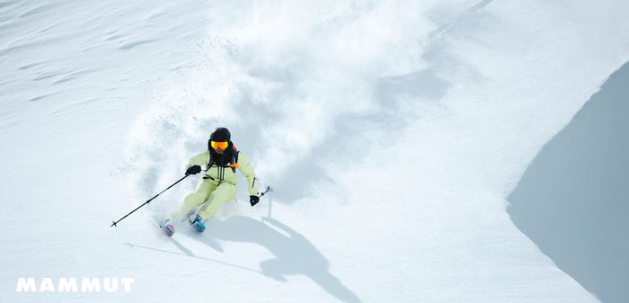 Skibekleidung aus leuchtend gelb-grünem Stoff mit schwarzen Akzenten. Skifahrer verwendet Stöcke auf weißem Schnee, der eine Pulverschnee-Sprühwolke erzeugt. Schwarzer Helm und Skibrille.