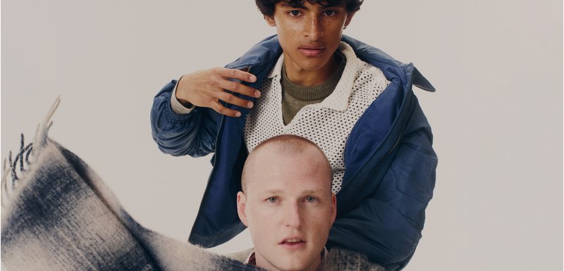 Deux personnes placées ensemble ; l'une a des cheveux bouclés et porte une veste en duvet bleue, l'autre est chauve avec un pull en tricot et un pantalon à carreaux.