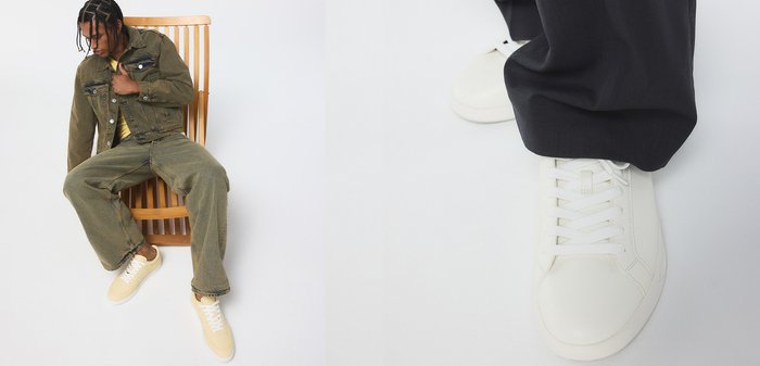 Man met gevlochten haar zit op een houten lattenstoel, draagt een groene spijkerjasje, wijde jeans, een geel hemd en beige sneakers.