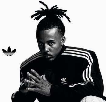 Image en noir et blanc d'un homme portant une veste de survêtement Adidas noire avec des rayures blanches. Il a des dreadlocks et les mains jointes devant lui.