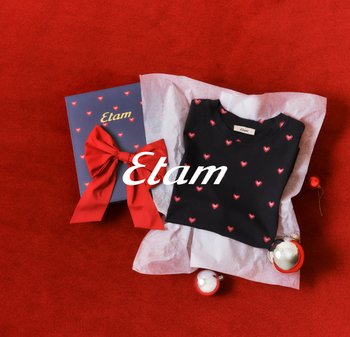 Czarny t-shirt z czerwonymi wzorami serc, leżący na pogniecionym papierze bibułkowym obok pudełka prezentowego z dużą czerwoną kokardą. Czerwone tło.