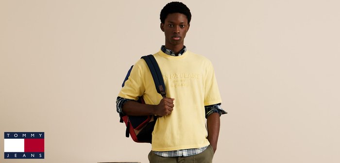 Camisa amarilla claro de manga corta con bordado texturizado, llevada sobre una camisa de cuadros azul y blanca. Una mochila azul marino y roja está colgada del hombro.