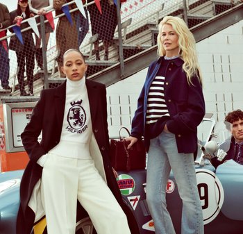 Deux femmes se tiennent à côté d'une voiture vintage. L'une porte un manteau noir sur un pull blanc avec un logo et un pantalon blanc. L'autre arbore une veste marine sur une chemise rayée et un jean évasé, tenant un sac à main bourgogne.