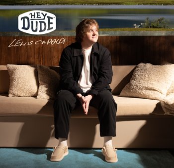 Scarpe beige slip-on con una superficie testurizzata, punta leggermente arrotondata e dettagli minimi. Indossate con pantaloni neri e calze bianche su un divano chiaro.