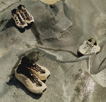 Trois paires de chaussures de randonnée beige et marron posées sur des surfaces rocheuses grises inégales en plein air sous la lumière du soleil.