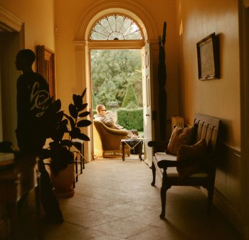 Couloir intérieur avec un sol en bois clair, comprenant un fauteuil vintage, un banc et une porte donnant sur un jardin. Éclairage doux et chaleureux.