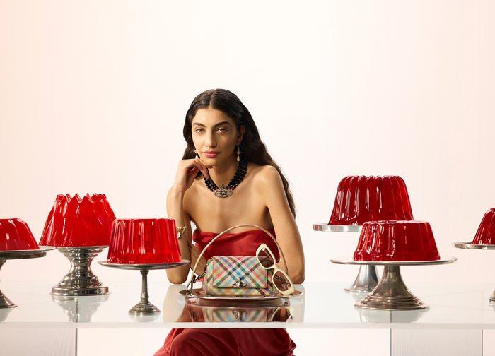 Γυναίκα με κόκκινο φόρεμα χωρίς τιράντες που φοράει μαύρο κολιέ, καθισμένη σε γυάλινο τραπέζι με κόκκινα γλυκίσματα ζελατίνης και πολύχρωμη τσάντα με λευκά γυαλιά ηλίου.