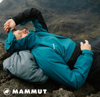 Tiefblaue wasserdichte Jacke mit schwarzen Akzenten, verstellbarer Kapuze und Reißverschlusstaschen, kombiniert mit einem grauen strukturierten Rucksack, der auf felsigem Terrain liegt.