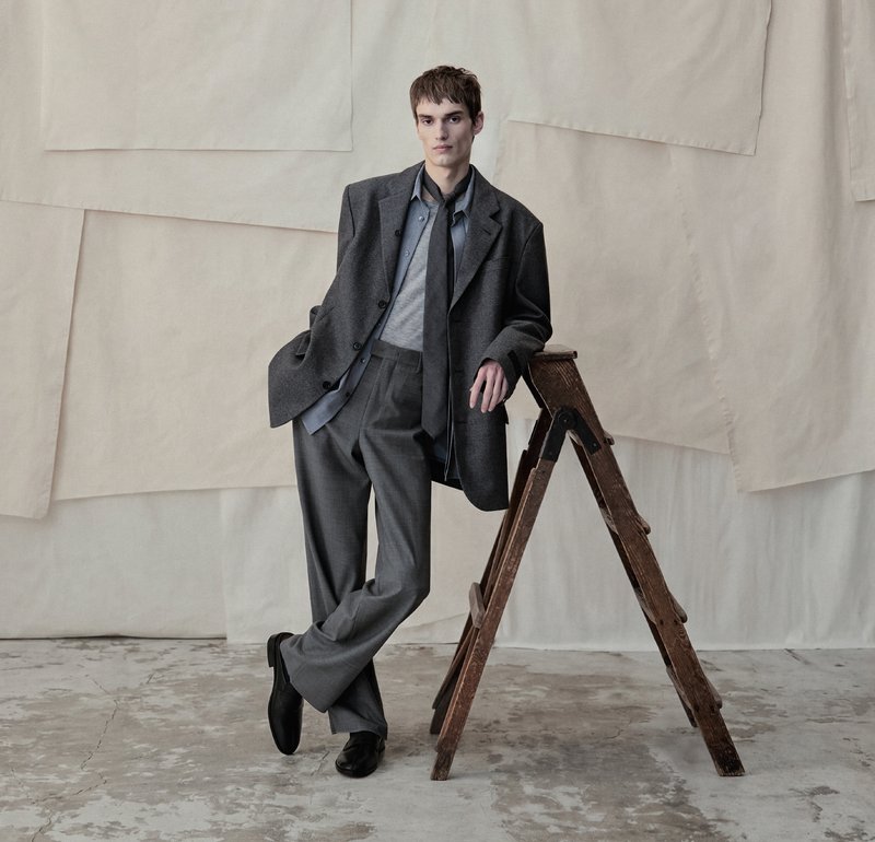Jeune homme en costume gris oversize et chaussures noires appuyé sur une échelle en bois dans un studio avec un fond en tissu beige.