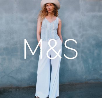 Kobieta w pasiastym bezrękawniku, szerokich białych spodniach i kapeluszu z szerokim rondem, stojąca na tle szarej, teksturowanej ściany.