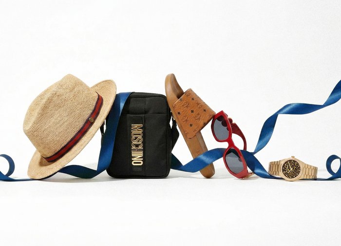 Chapeau de paille, sac noir Moschino, chaussure de créateur couleur sable, lunettes de soleil rouges et montre-bracelet en or reliés par un ruban bleu fluide sur fond blanc.