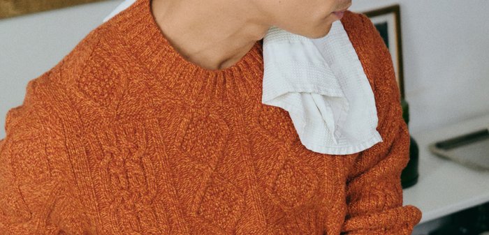 Pull orange en maille torsadée avec poignets et ourlet côtelés. Personne tenant un verre à vin transparent. Plan de travail de cuisine avec divers ustensiles et ingrédients.