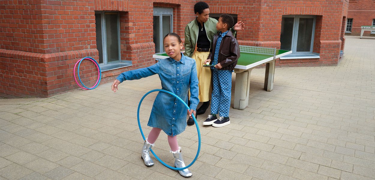 Enfant en robe en denim fait tourner un hula hoop bleu tandis que deux autres, dont un tenant des raquettes de tennis de table, se tiennent près d'une table de ping pong devant un bâtiment en briques.