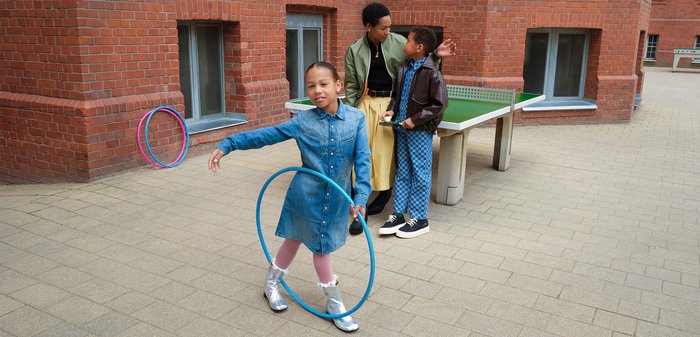 Enfant en robe en denim fait tourner un hula hoop bleu tandis que deux autres, dont un tenant des raquettes de tennis de table, se tiennent près d'une table de ping pong devant un bâtiment en briques.