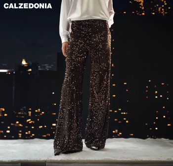 Personne portant un pantalon à paillettes marron taille haute à jambes larges et un chemisier blanc se tient devant un paysage urbain sombre la nuit.