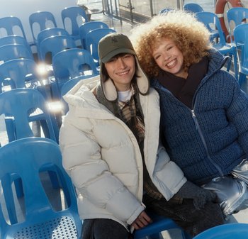 Δύο άτομα κάθονται σε μπλε πλαστικές καρέκλες. Ο ένας φοράει λευκό μπουφάν puffer, ο άλλος ένα μπλε υφασμάτινο puffer. Και οι δύο φορούν σκούρα τζιν και έχουν casual στυλ.