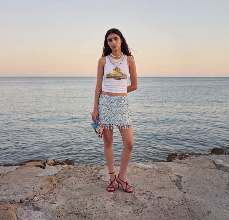 Femeie cu top alb și fustă cu model stă pe o țărm stâncos cu mare liniștită și cer pastelat în fundal la apus.
