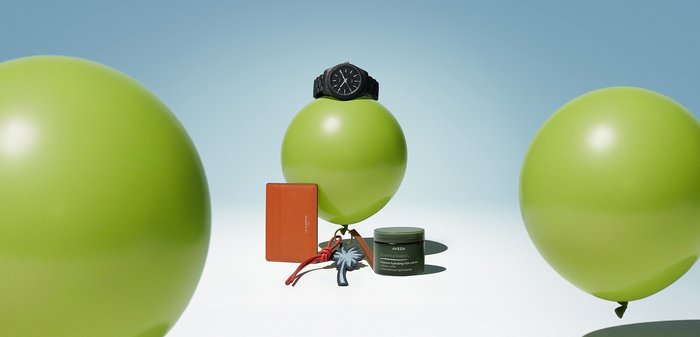 Drei grüne Luftballons mit Luxusartikeln balanciert, darunter eine schwarze Armbanduhr auf dem mittleren Ballon, ein Hautpflegeglas, ein orangefarbener Kartenhalter und ein Schlüsselanhänger.