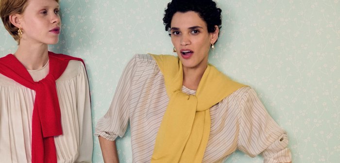 To kvinder poserer mod en blegmønstret væg, iført lyse bluser, blå jeans og trøjer lagt over skuldrene i rød og gul.