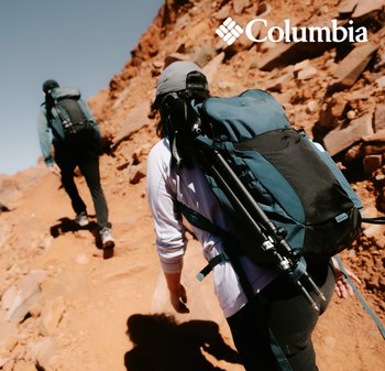 Deux randonneurs sur un sentier rocailleux. L'un porte un sac à dos noir et bleu sarcelle avec des sangles externes pour les bâtons de randonnée. Tous deux sont vêtus de vêtements de plein air.