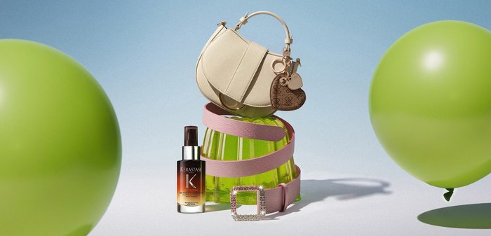 Bolso beige con cinturón rosa alrededor de un jarrón de cristal verde, junto a una botella marrón de suero para el cabello, rodeado de dos globos verdes grandes sobre fondo azul.