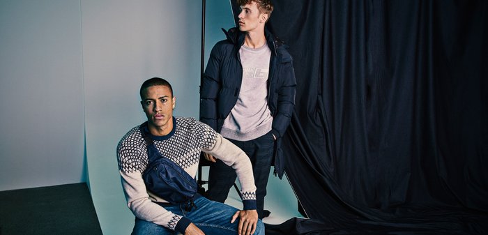 Due modelli mostrano abbigliamento esterno: uno indossa un maglione a fantasia con accenti scuri, l'altro una giacca a vento nera sopra una felpa grigia con colletto rotondo.