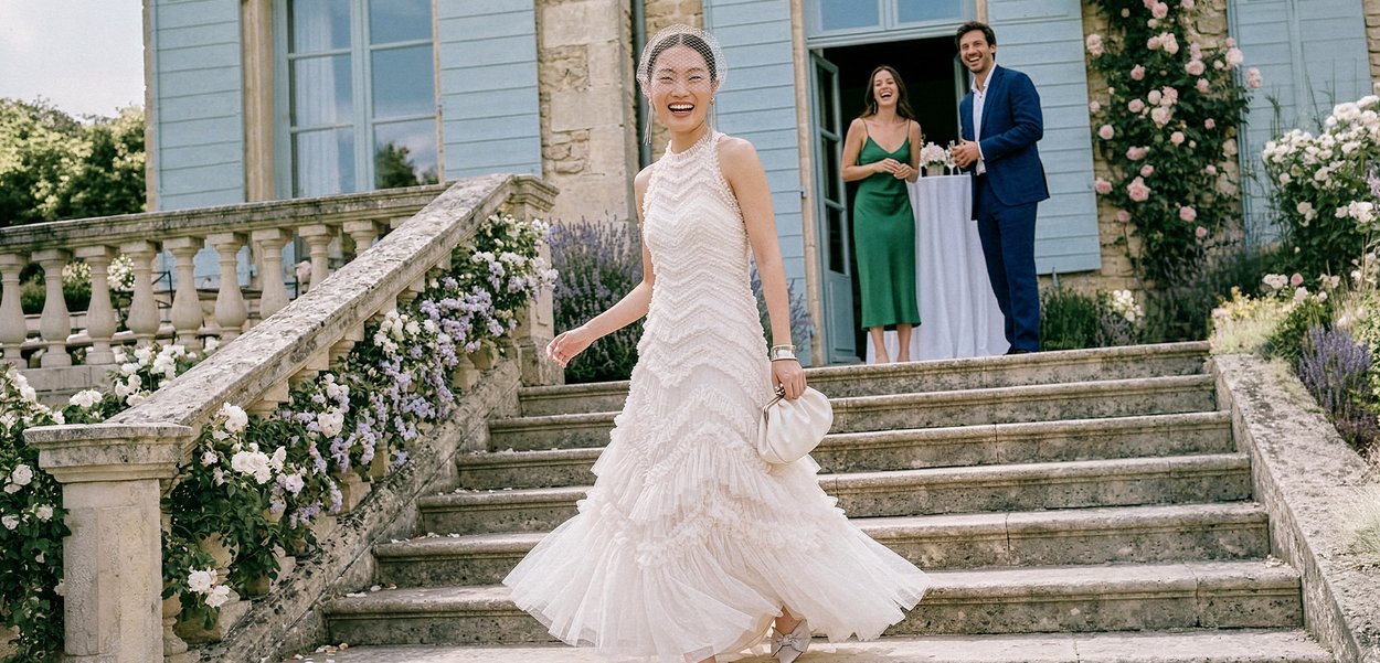 Novia con vestido de novia blanco girando bajando unos escalones de piedra bordeados de flores, con dos invitados sonriendo y observando cerca de la entrada de un edificio azul.