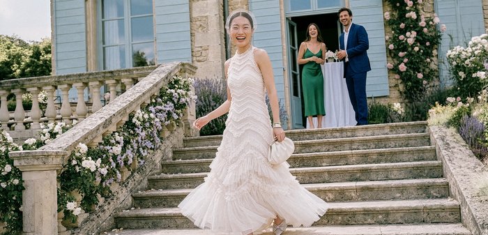 La mariée en robe blanche tourne en descendant des marches en pierre bordées de fleurs, tandis que deux invités sourient et regardent près de l'entrée d'un bâtiment bleu.