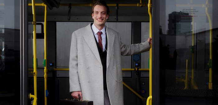 Manteau en laine gris, gilet noir, cravate rouge à motifs. Porte une mallette noire, debout à la porte d'un bus, avec des barres de maintien jaunes visibles.
