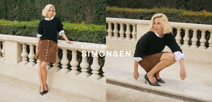 Blondínka v čiernom svetri, bielej košeli, hnedej sukni a čiernych lodičkách drepe na kamennom okraji s zeleňou v pozadí.