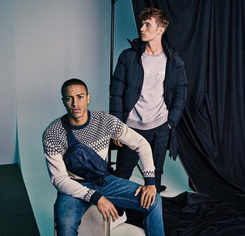 Due modelli maschili mostrano abbigliamento. Uno indossa un maglione chiaro a motivi, jeans blu e una borsa a tracolla scura; l'altro indossa un giubbotto imbottito nero e un top grigio.