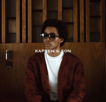 Mladý muž se slunečními brýlemi, bílou košilí a vzorovaným bordó kardiganem sedí s opřením o dřevěné panely.