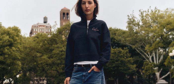 Young woman wearing black Calvin Klein pullover and wide-leg jeans stands by stone benches in park holding black handbag.