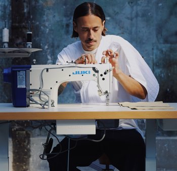 Symaskine JUKI med hvidt og blåt kabinet, justerbar stinglængde og trådspoler på stativer, betjent af en person i en hvid skjorte.