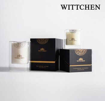 Dwie świece w szklanych pojemnikach, jedna przezroczysta z złotym wzorem, oraz dwa czarne pudełka z złotymi akcentami oznaczone "Wyważony Urok" i "Euforia Szampana."