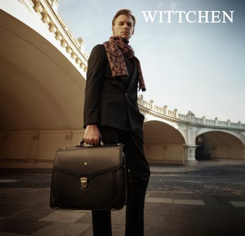 Czarna skórzana teczka na dokumenty z metalowym zapięciem, trzymana przez mężczyznę w ciemnym garniturze i wzorzystym brązowym szaliku, na tle architektonicznym.