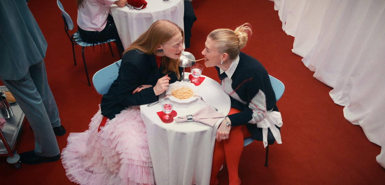 Divas sievietes dalās ar vienu spageti šķēli pāri galdam, uz kura ir šķīvis ar spageti, ūdens glāzes un balti galdauti uz sarkana paklāja.