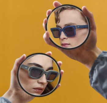 Gafas de sol con montura gruesa, una azul y otra gris, cada una con lentes de tonalidad oscura. Reflexiones visibles en un espejo circular de mano.