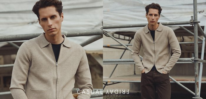 Beige gestrickte Cardigan mit Reißverschluss und Kragen, getragen über einem schwarzen Hemd. Das Modell steht vor einem Baustellenhintergrund, die Hände in den Taschen.