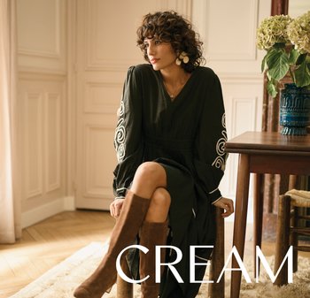 Frau mit lockigem Haar in einem schwarzen Kleid mit weißer Stickerei sitzt auf einem Holzstuhl, trägt braune kniehohe Stiefel neben einem Tisch mit einer Vase.