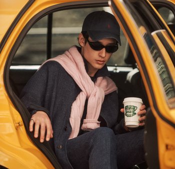Persona con gafas de sol, gorra azul marino y bufanda, sentada en un taxi amarillo sosteniendo una taza de café.