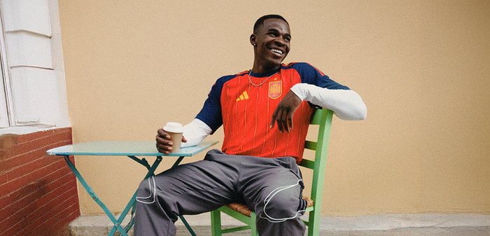 Maillot de sport rouge et marine avec des rayures verticales et un logo, associé à un pantalon gris. Chaise pliante verte et tasse de café sur une table.