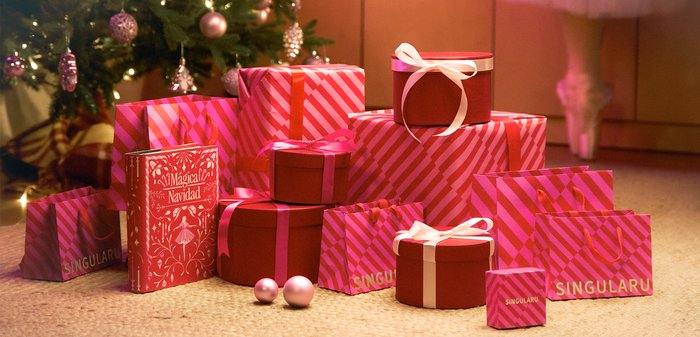 Regali avvolti colorati di varie dimensioni, principalmente rosa con accenti bianchi e motivi a righe, accanto a un libro rosso decorativo e ornamenti.