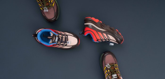 Quatre chaussures de sport posées sur une surface sombre, deux en rouge et beige et deux en violet et rose avec des lacets maintenus par des attaches plastiques.