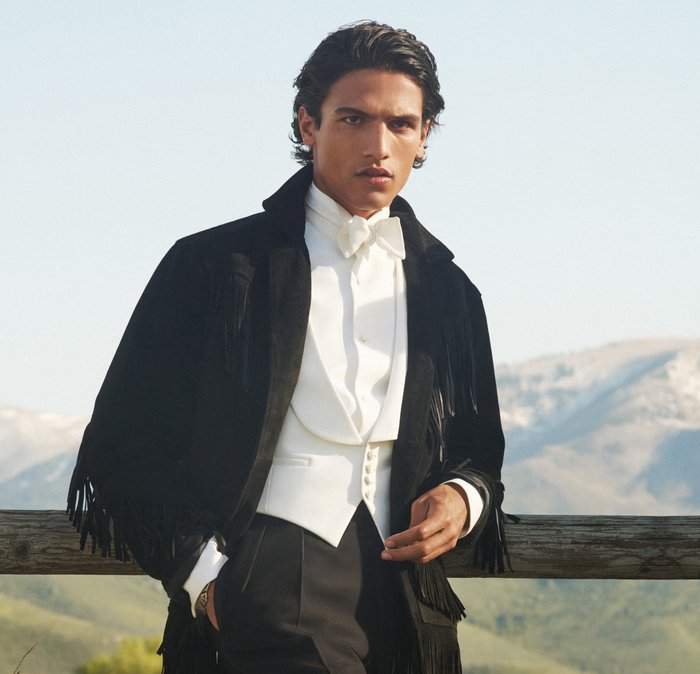 Man wearing a white tuxedo shirt and bow tie with a black fringed jacket, standing by a wooden fence with mountains in the background.