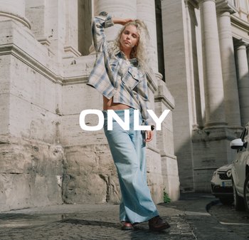 Jonge vrouw in een geruite blouse en ruime spijkerbroek die zelfverzekerd poseert voor grote stenen zuilen met een geparkeerde auto in de buurt, met de tekst "ONLY" eroverheen.