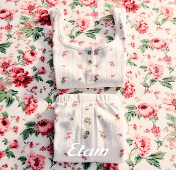 Witte geribbelde top en korte broek van stof met kleine roze roosjes en knopen, gelegd op een bijpassende bloemenachtergrond.